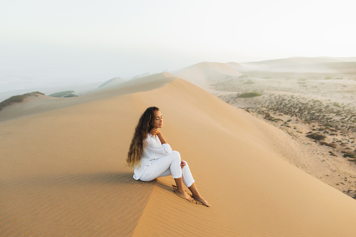 Beautiful brunette woman in white clothes relaxing on top of sand dune in Morocco Sahara desert. Mist in morning before sunrise. Harmony with nature.