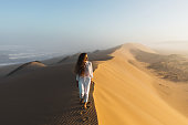 Woman in white clothes walking by top of huge sand dune near ocean coast in Morocco. Beautiful warm sun light and mist in morning. Sahara desert.