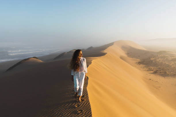 woman in white clothes walking by top of huge sand dune near ocean coast in morocco. beautiful warm sun light and mist in morning. sahara desert. - fog desert arabia sunset imagens e fotografias de stock