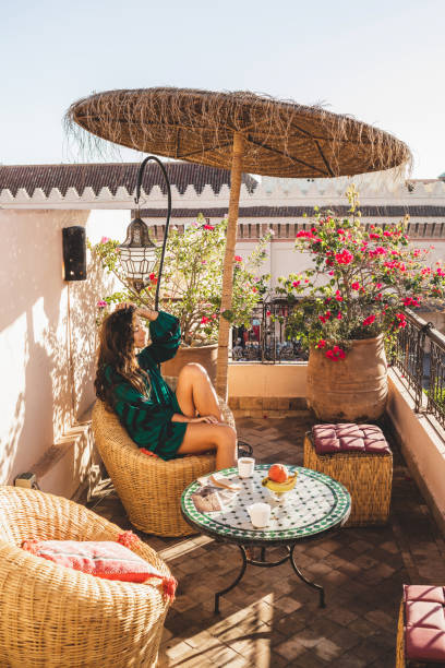 Woman enjoying breakfast on beautiful lounge hotel terrace in downtown of Marrakech at sunrise. Moroccan design with vintage elements - metal lantern, wicker chair, round table, and umbrella. Woman enjoying breakfast on beautiful lounge hotel terrace in downtown of Marrakech at sunrise. Moroccan design with vintage elements - metal lantern, wicker chair, round table, and umbrella. moroccan girl stock pictures, royalty-free photos & images