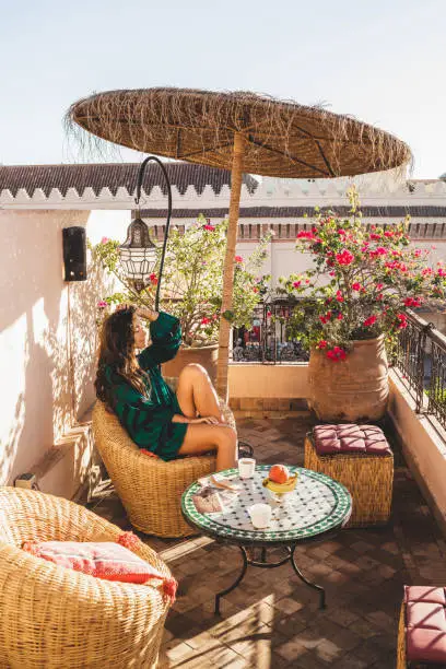 Photo of Woman enjoying breakfast on beautiful lounge hotel terrace in downtown of Marrakech at sunrise. Moroccan design with vintage elements - metal lantern, wicker chair, round table, and umbrella.