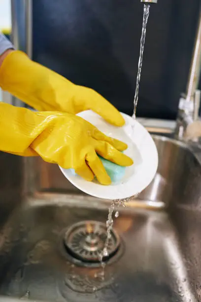 Close-up image of housewife in rubber gloves washing dishes and rinsing with tap water