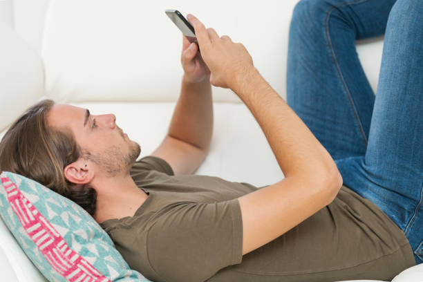 istock Close up of a handsome young man using his phone while lying on a sofa. Relaxation and communication concept. 1264937120
