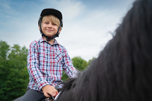 Happy smiling young blonde boy riding his black horse, smiling looking over to the camera. Natural outdoor horseback riding portrait. Real People Riding Club Horseback Riding Lifestyle