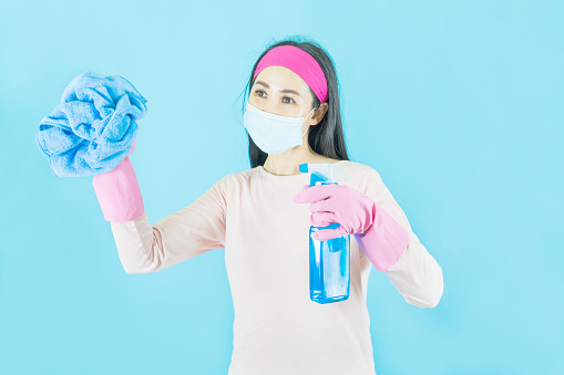 Joven mujer asiática ama de casa en guantes rosas llevaba máscara de la cara, botella de spray en la mano está limpia aislado sobre fondo azul, tareas domésticas y concepto del hogar. photo