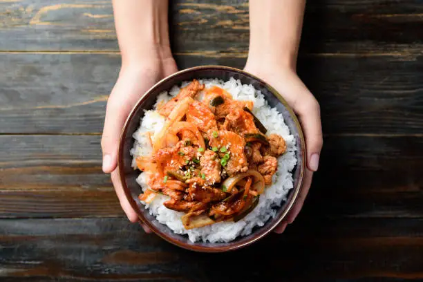 Korean food, Stir fried kimchi with pork on cooked rice in a bowl holding by hand on wooden background, Top view