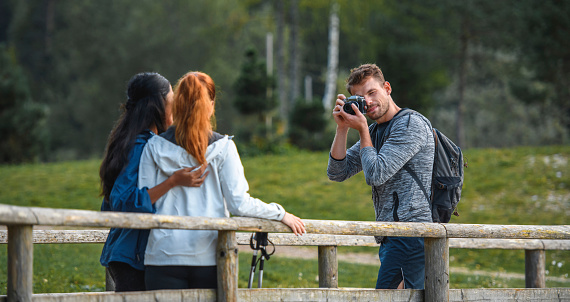 Smiling male hiker in early 30s taking a break to photograph female friends in their 20s on pedestrian bridge in northwestern Slovenia.