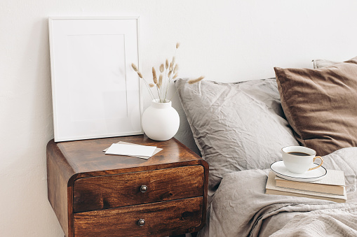 Portrait white frame mockup on retro wooden bedside table. Modern white ceramic vase, dry Lagurus ovatus grass. Cup of coffee and books in bed, beige linen pillows in bedroom. Scandinavian interior.