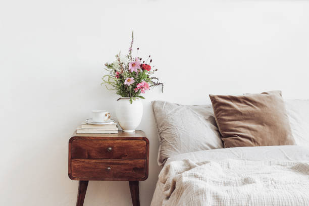 xícara de café e livros em mesa de cabeceira de madeira retrô. vaso de cerâmica branco rústico com buquê de cocmos rosa e flores de zínia. linho bege e travesseiros de veludo na cama. interior escandinavo, quarto. - bedroom indoors wall bedding - fotografias e filmes do acervo