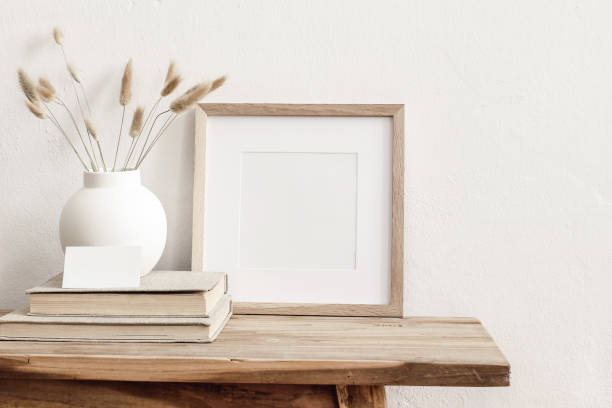 istock Square wooden frame mockup on vintage bench, table. Modern white ceramic vase with dry Lagurus ovatus grass, books and busines card. White wall background. Scandinavian interior. 1264970165