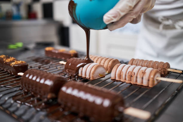 Dessert chef covering a cake biscuit on a popsickle with a melted black chocolate on a metal net tray
