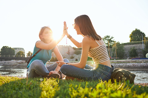 Cheerful teenage female friends using their free time to relax at the quayside while sitting on a grass in sunset and greeting each other with a high five