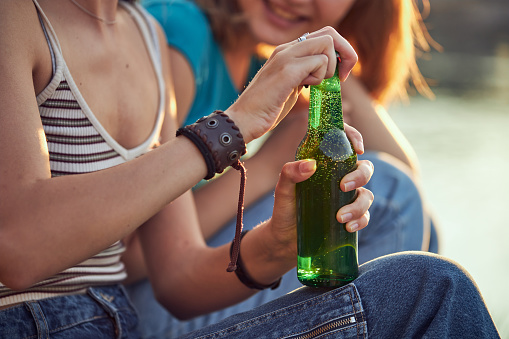 Cheerful female friends sitting on a quayside and spending time together while one of a woman opening a beer bottle