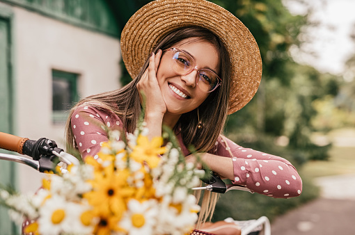 Optimistic young female in stylish hat and glasses smiling and looking at camera while sitting on bike with bouquet of fresh flowers, on summer day in countryside
