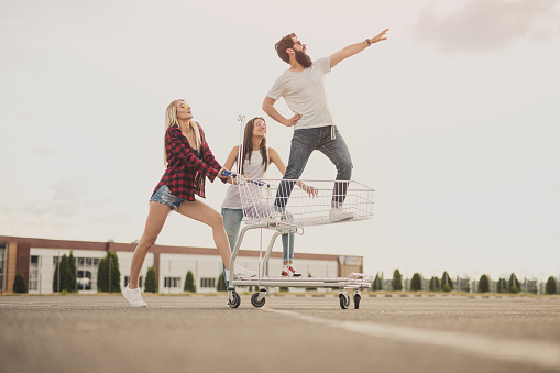 Full body positive bearded guy pointing away while standing in shopping cart near female friends on summer day on parking lot