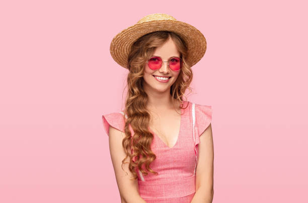Happy young female in summer hat and sunglasses smiling and looking at camera against pink background