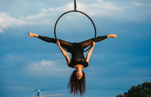 Woman aerialist performs acrobatic element split in hanging aerial hoop against background of blue sky and white clouds.
