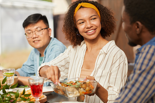 Portrait of smiling African-American woman sharing food while enjoying dinner with friends and family outdoors at Summer party, copy space
