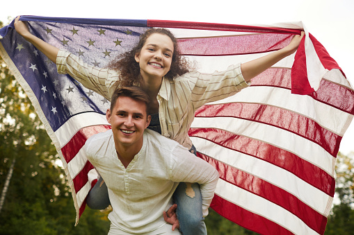 Low angle portrait of carefree young couple looking at camera while waving American flag outdoors, copy space