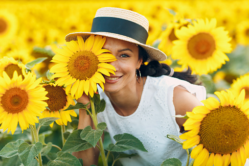 Beautiful smiling young woman in a hat with flower on her eye and face on a field of sunflowers.