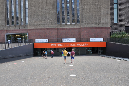 Tate Modern art gallery and museum. Turbine Hall entrance with orange Welcome sign.  Tourists including two men walking towards the entrance near round coronavirus social distancing signs on the ground. Bright summers day. River Thames South Bank, London, United Kingdom, August 2, 2020