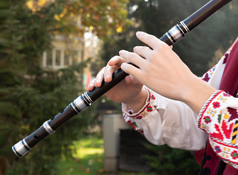 Hands of young musician play on wooden flute. Man in Bulgarian national costume plays on long pipe. Kaval is Balkan folk traditional woodwind musical instrument.