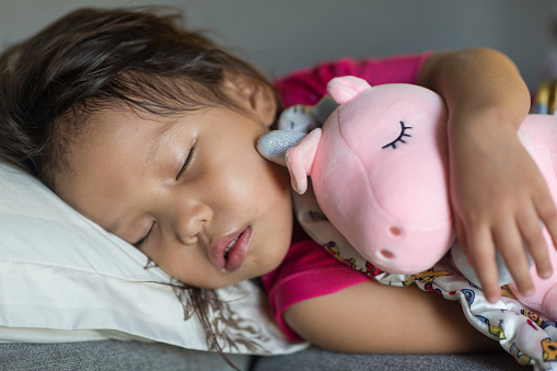 Adorable young female child asleep on a comfortable pillow and bed while cuddling with a pink stuffed animal.