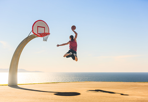 Young teen athlete playing basketball and elevating up in the air towards the basket at a outdoor court with the beautiful ocean view background at sunset. Sports boost and energy.