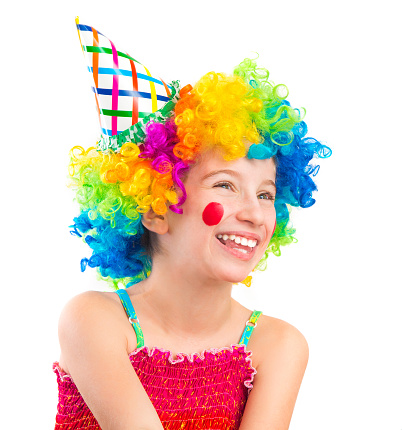 This is a close-up image of a boy who is wearing a colorful clown wig, a jumbo bow tie, and a big red nose. The child is cheerfully looking towards the camera with party flags and balloons in the background.