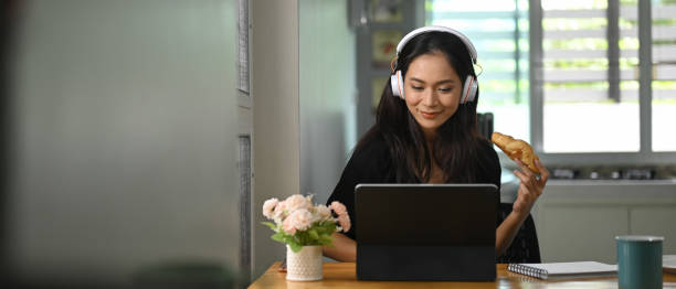 a beautiful woman is eating a croissant while using a computer tablet at the wooden working desk. - food staple audio imagens e fotografias de stock