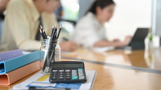 A calculator is putting on the wooden table surrounded by various office equipment over employees as a background.