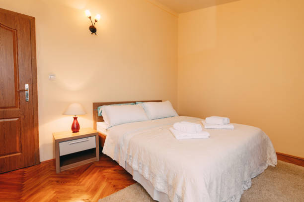 istock Large queen size bed with pillows and fluffy towels on the bedspread in a spacious room with reading and wall lamps and doormat. 1265038310