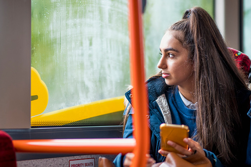 Mixed race teenage girl commuting to school holding her smartphone and looking out of the window.