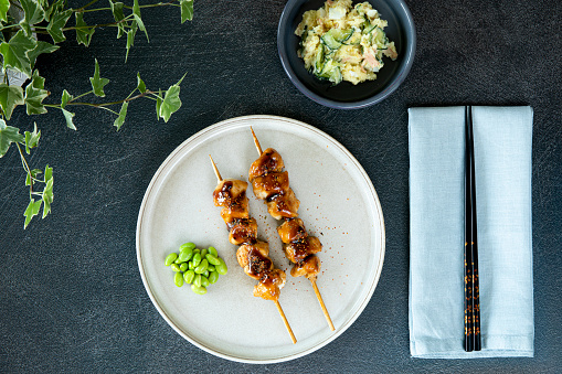 Plate of 2 chicken yakitori on skewers, edamame beans on the side, bowl of Japanese potato salad, on dark metal background, pair of black chopsticks, pale pastel blue napkin, pot plant in the corner