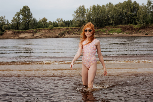 Red-haired girl in a swimsuit stands in the water on a hot summer day.