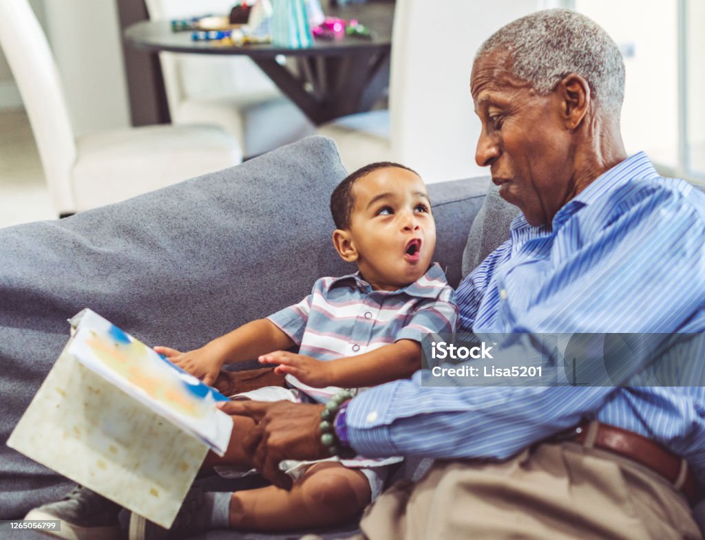 African American grandchild and grandfather read a book together at home Adorable toddler with his grandfather relaxing at home reading a book together Grandparent Stock Photo