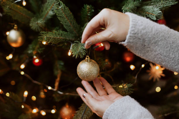 Woman decorating christmas tree with shiny golden bauble closeup. Preparation for christmas time. Modern glitter ornament in hands on background of festive tree in lights. Happy holidays stock photo