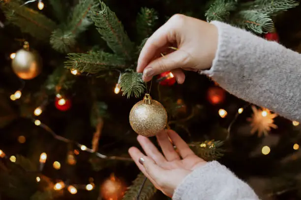 Photo of Woman decorating christmas tree with shiny golden bauble closeup. Preparation for christmas time. Modern glitter ornament in hands on background of festive tree in lights. Happy holidays