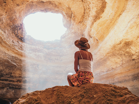Rear view of woman sitting on top of rock inside sea cave in Portugal, sunbeam passing through hole illuminating the while place