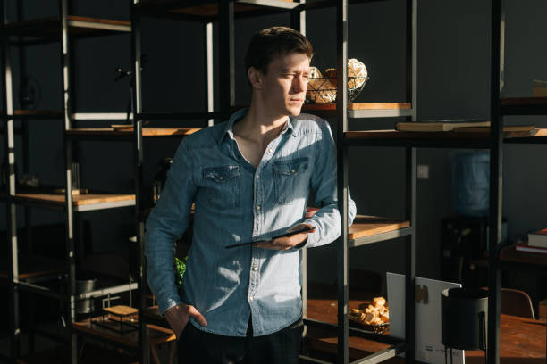 istock Handsome young man using digital tablet pc while standing near shelves in office room. 1265074813