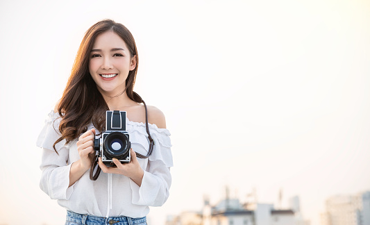 Portrait of beautiful asian woman photographer in fashion look taking photo. Pretty cool young woman model with retro film camera curly hair outdoors over rooftop sky with copy space banner