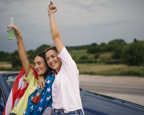 Two young happy Americans having fun in front their cabriolet outdoors.They are toasting together and wear American flag.Celebrate fourth of July.