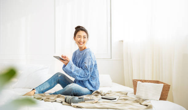 asiatische frau mit lächeln verwenden tablet-smartphone in blauen winterpullover arbeit nach hause, portrait junge schönheit asia mädchen entspannen im schlafzimmer. technologie-menschen verandern digitales online-marktbanner - lifestyles female fashion model beautiful stock-fotos und bilder