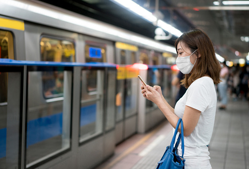Asian woman texting at the metro station on her cell phone wearing a facemask to avoid an infectious disease