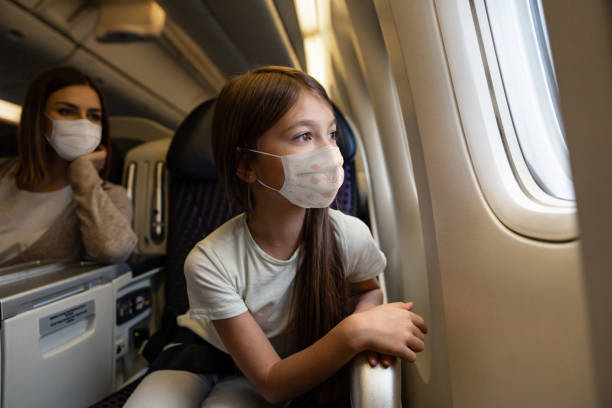 Happy girl traveling by plane wearing a facemask Portrait of a happy girl traveling by plane wearing a facemask and looking through the window - travel concepts arrival departure board photos stock pictures, royalty-free photos & images