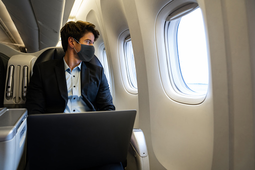 Business man traveling by plane wearing a facemask and using his laptop while looking through the window â COVID-19 pandemic