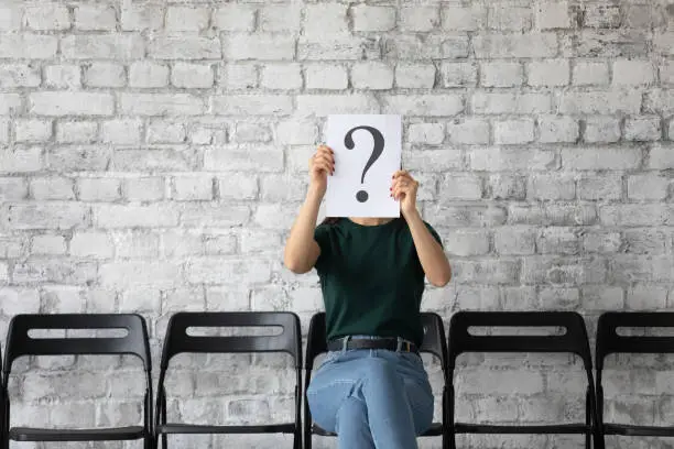 Young woman candidate hiding face behind sheet with question mark, sitting on chair in empty office hall, unemployed applicant seeker waiting for job interview, employment and hiring concept