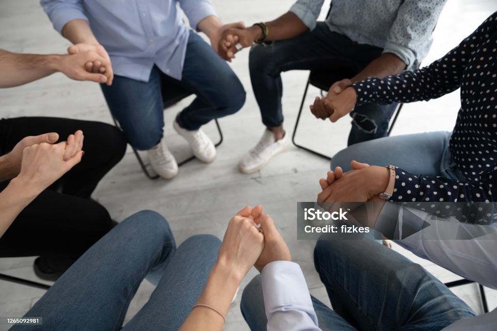 Close up diverse people sitting in circle at psychological session Close up diverse people sitting on chairs in circle at group training counselling session, holding hands, psychological help and treatment concept, drug or alcohol addiction rehabilitation Empathy Stock Photo
