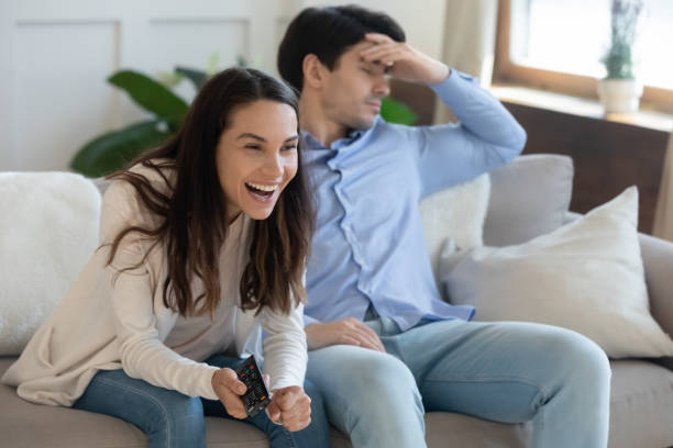 Couple disagree on channel choice or tv program. Unhappy sad bored young man sitting separately on cozy couch while smiling millennial woman using remote controller, watching favorite series on television, couple disagreement on channel choice. divergent movie review stock pictures, royalty-free photos & images