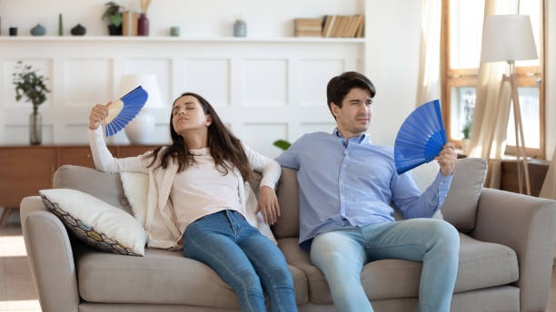 Stressed tired young family couple fanning fresh air. Stressed tired young family couple sitting together on sofa, fanning fresh cool air with paper appliances, feeling unwell at summertime, suffering from sunstroke or high temperature at home. air conditioner family stock pictures, royalty-free photos & images